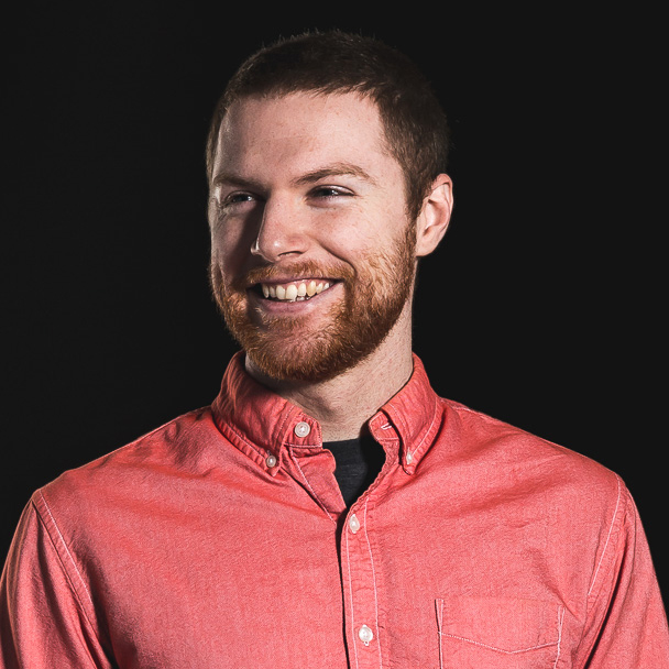 Glen smiling in a salmon button-up shirt on a black background.